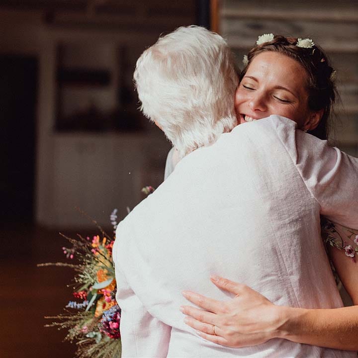 Bride with flowers in her hair hugs a wedding guest holding colourful wedding bouquet 