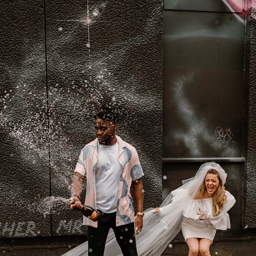 Groom in a pastel patterned shirt and Converse high tops popping champagne with his bride in a short wedding dress and long veil