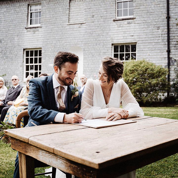 Groom in blue suit and gold patterned tie signs the register with the bride in Sassi Holford wedding gown