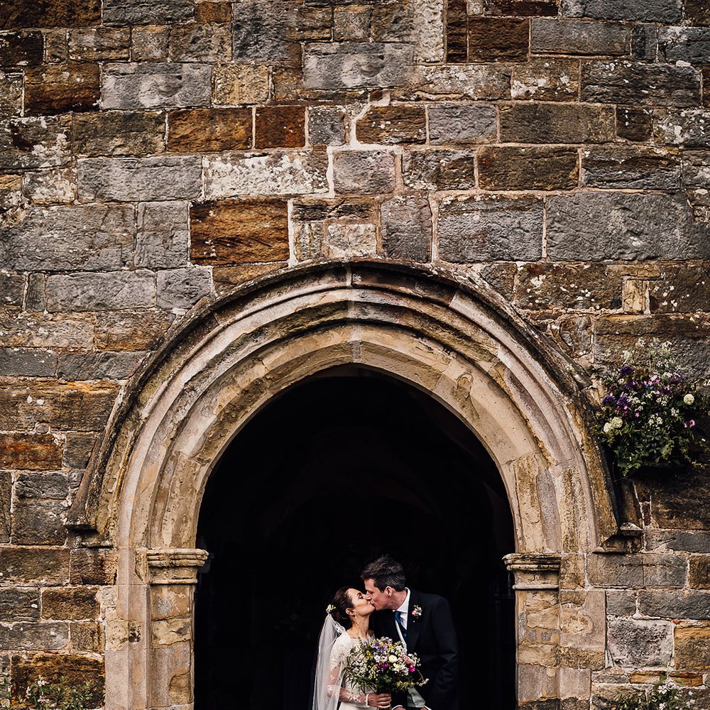 Groom in pinstripe trousers kisses bride in satin wedding dress under the arch entrance to the church with colourful bouquet 