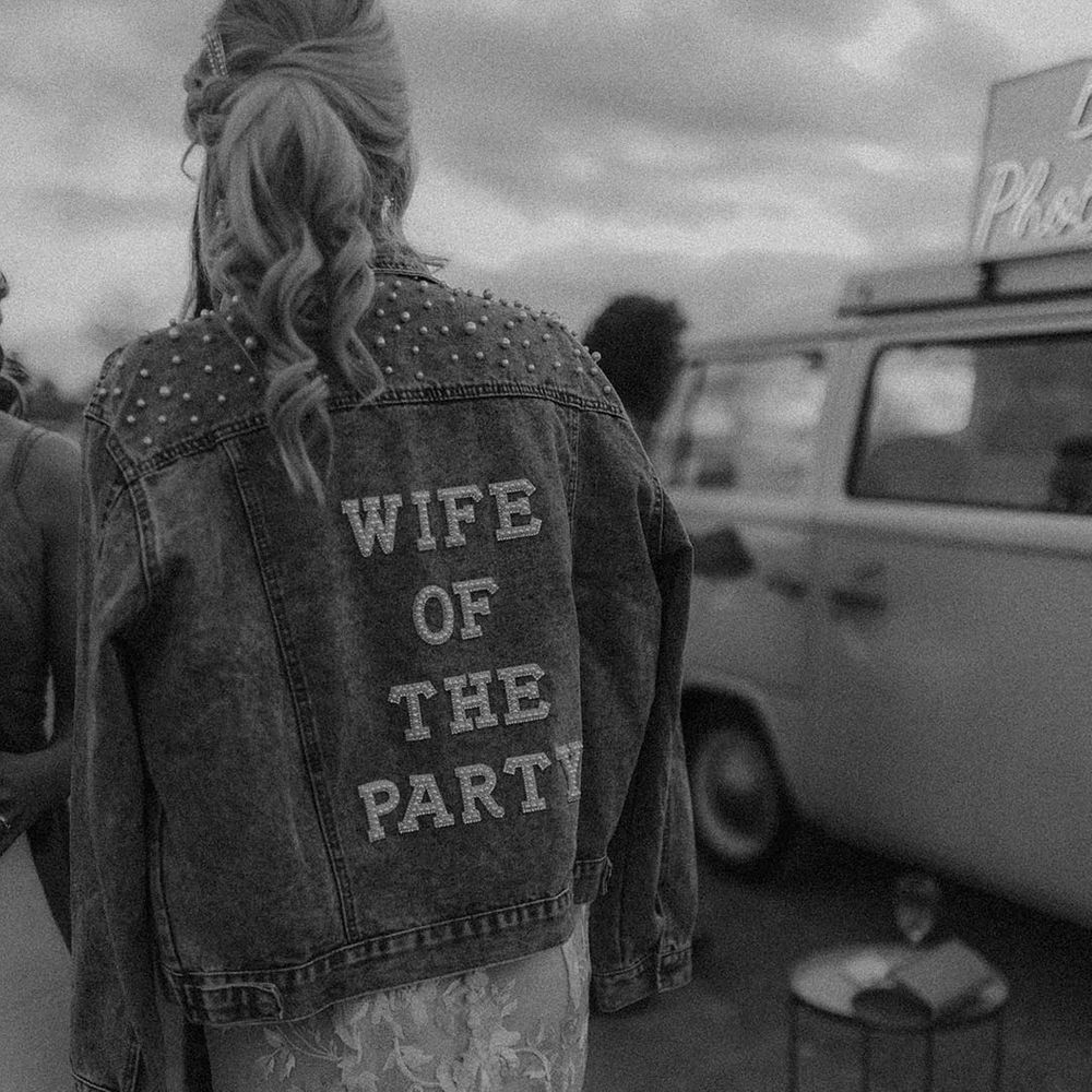 Bride wearing denim jacket embellished with pearls and back reading 'wife of the party' in the evening 