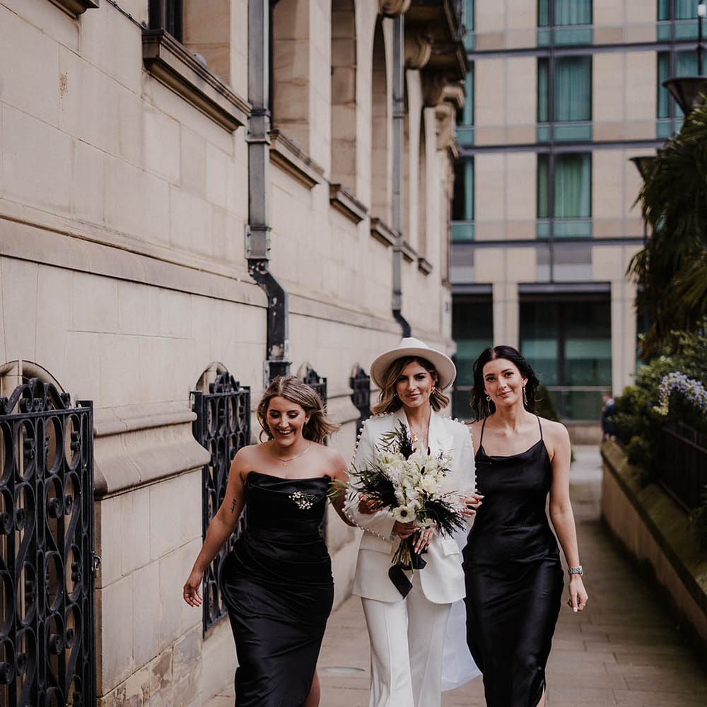 Bride wearing hat and pearl white trouser suit with bridesmaids in black satin dresses 