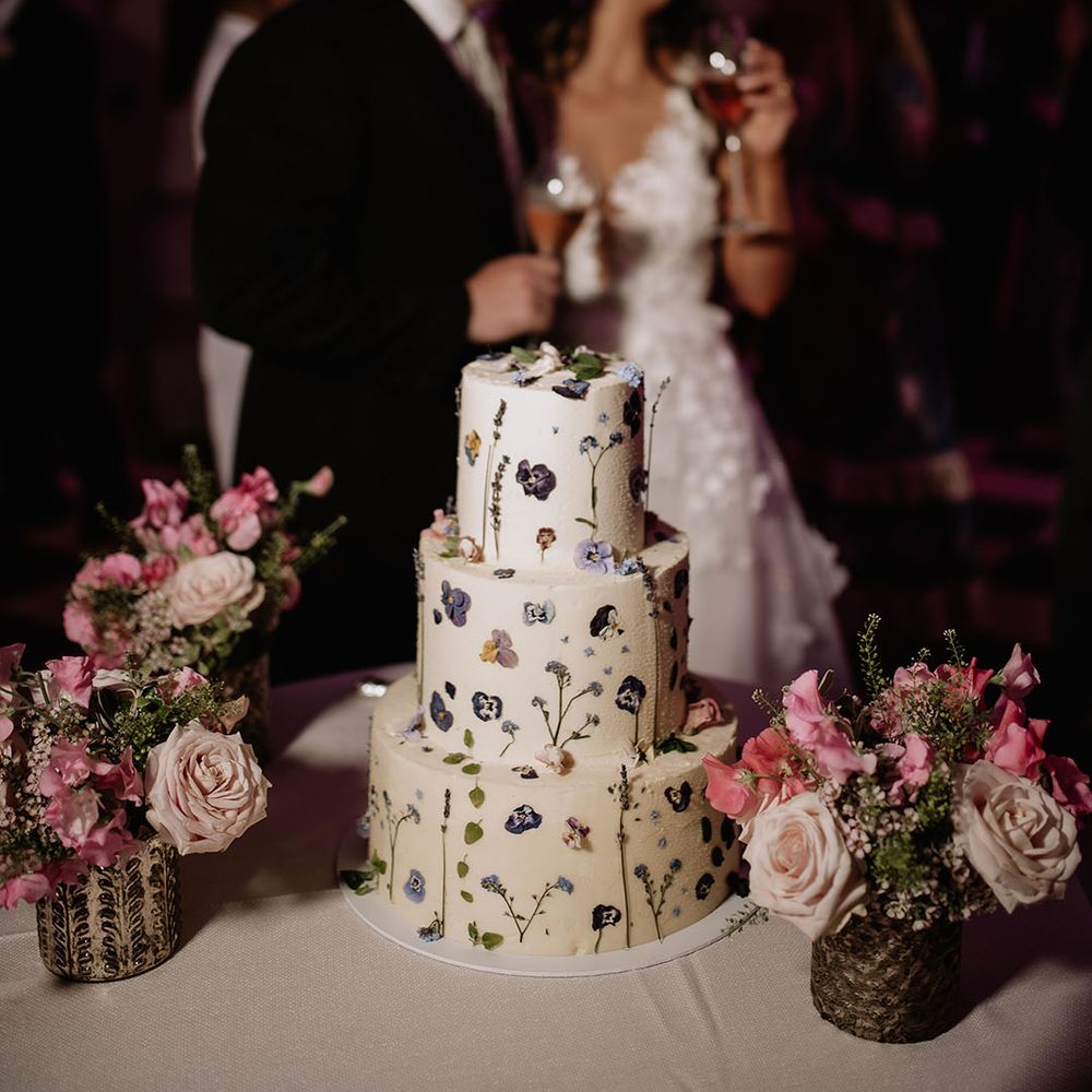 Three tier white frosted wedding cake with purple wildflowers and sprigs of lavender next to vases of pink flowers 