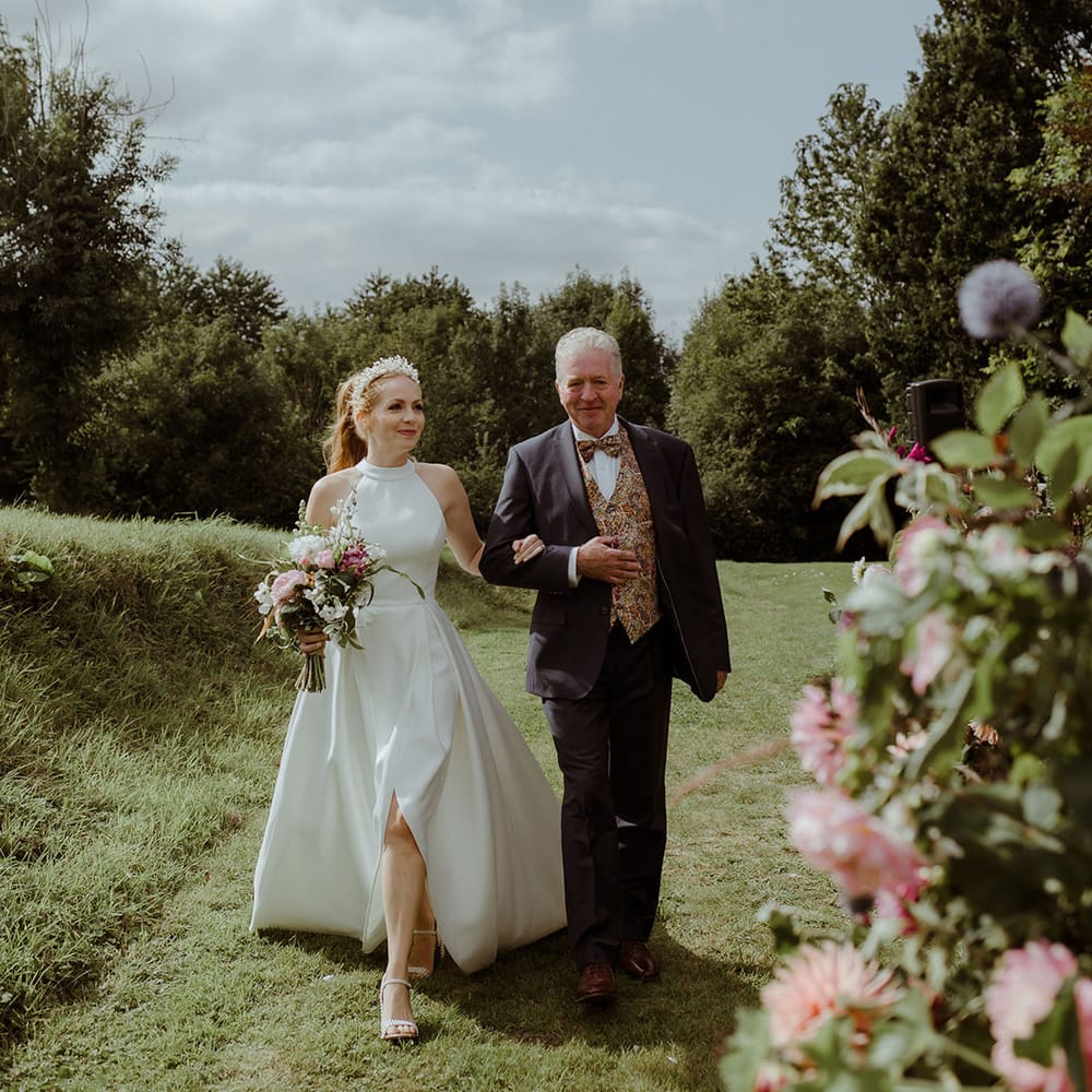 Bride in halter neck wedding dress wit pearl shoes is walked down the aisle by father of the bride in three piece suit with paisley tie and waistcoat 