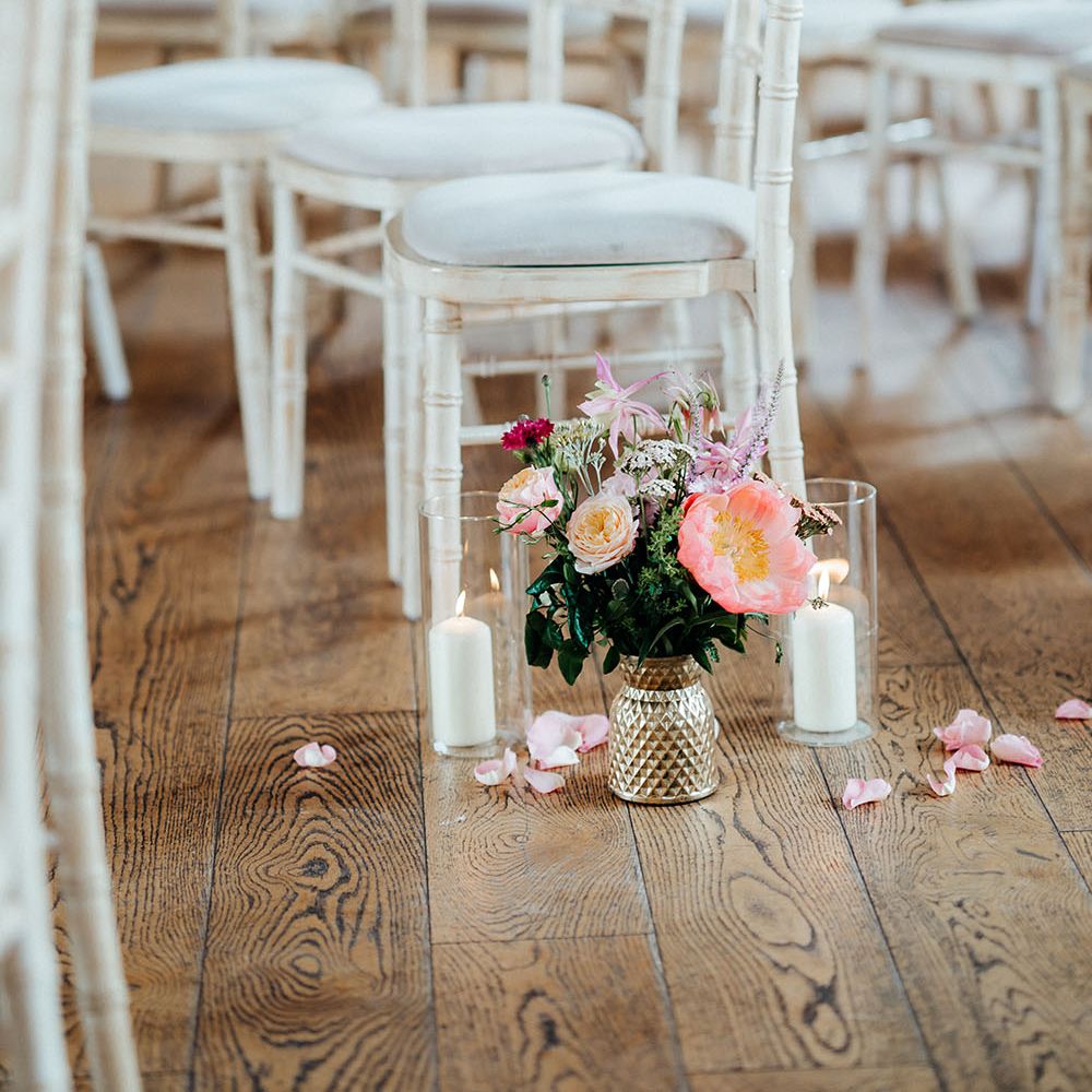 Bright floral bouquets in gold vases surrounded by petals and pillar candles in glass case beside white wicker chairs 