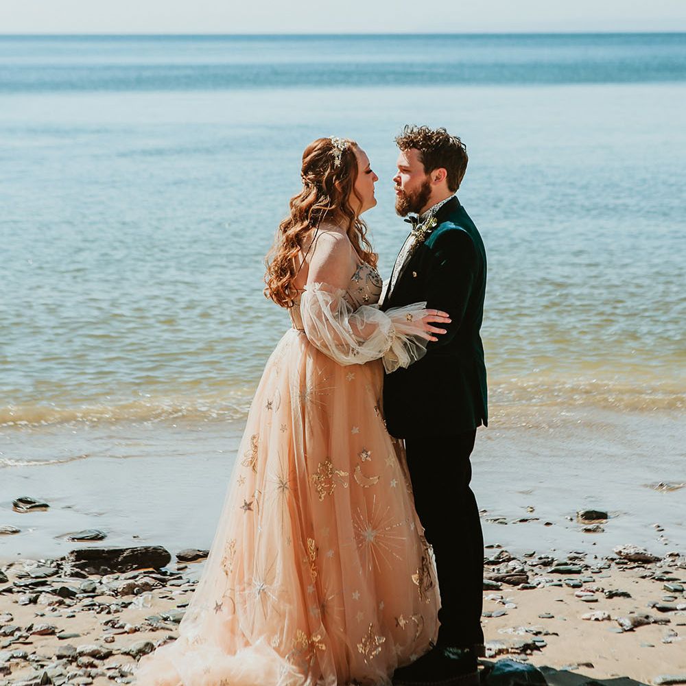 Bride wears off the shoulder starry wedding dress whilst stood with her groom in green velvet jacket on the beachfront in Cornwall 