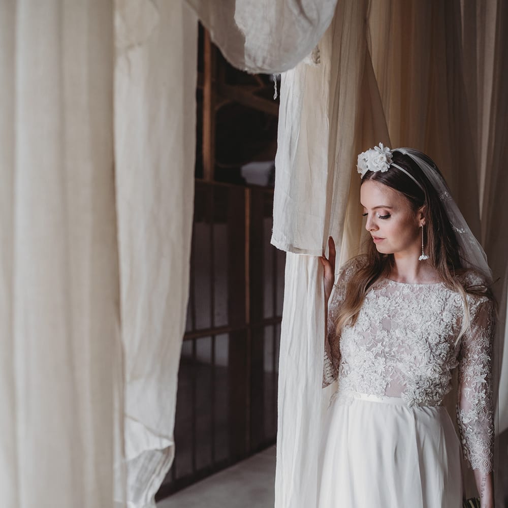 Bride stands in a lace wedding dress, embroidered veil, flower headband and earrings with a white flower bouquet