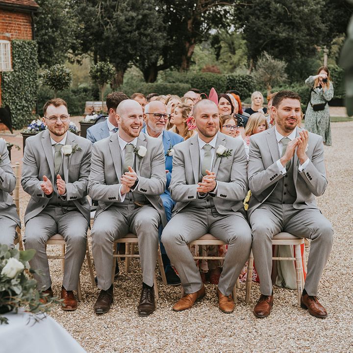 Groomsmen in matching light grey suits with sage green ties and white rose buttonholes applaud for the wedding 