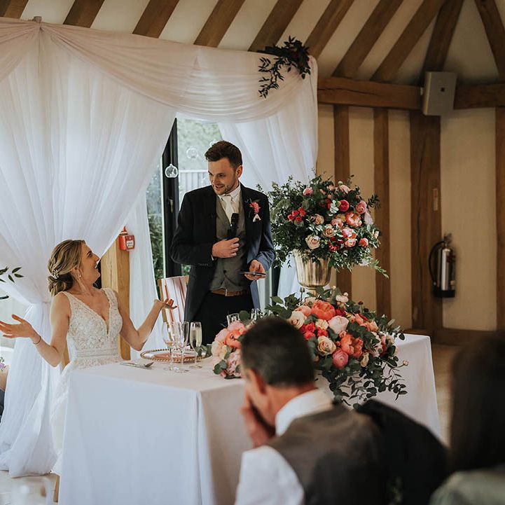 Groom stands to read out his speech as the bride sits at their sweetheart table decorated with pink flowers 