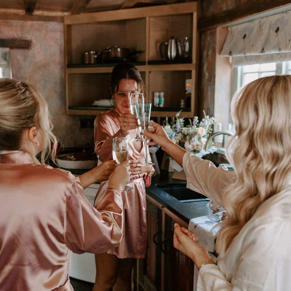 Bride in white satin robe shares a toast with the bridesmaids in pink satin robes as they get ready for the wedding 