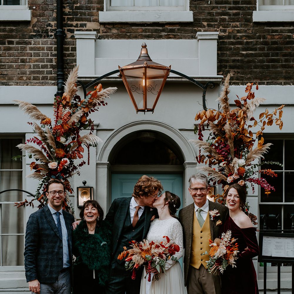 Bride in long sleeve lace vintage wedding dress and closed toe satin heels and groom in dark suit with orange tie and autumnal boutonniere kissing outside London townhouse with family of the bride and groom standing underneath large floral arch with roses, peonies, foliage and pampas grass