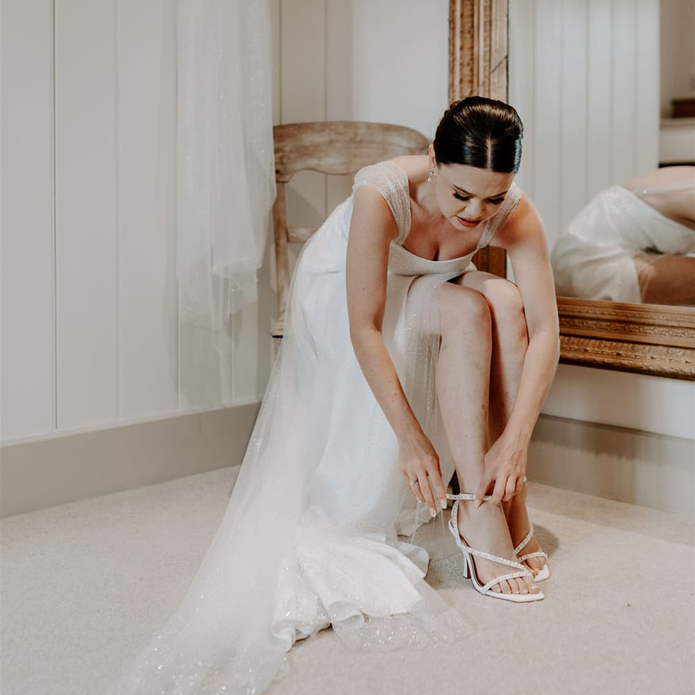 Bride wearing sparkly wedding dress puts on her white wedding shoes with DIY pearls 
