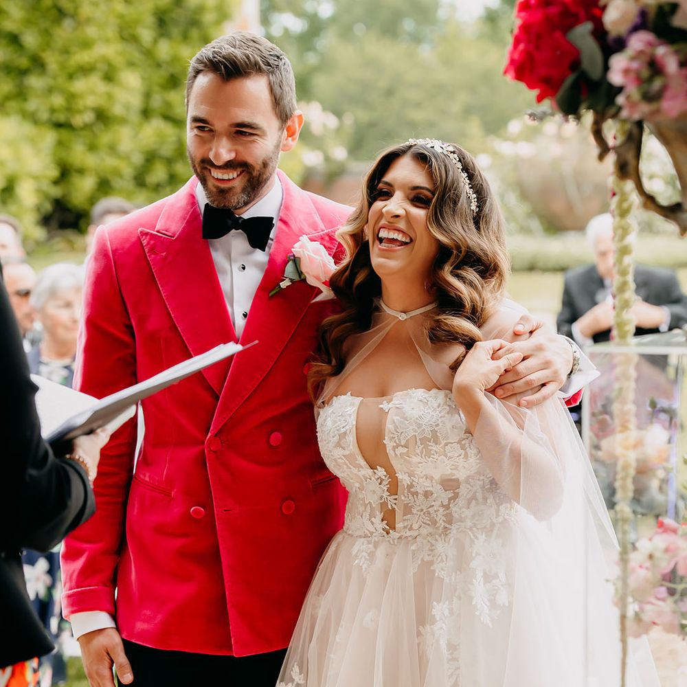Groom in a dark bright pink velvet jacket and light pink rose buttonhole has his arm around the bride in a tulle gown with sheer cape for their outdoor ceremony 