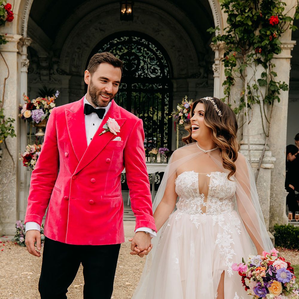 Groom in striking pink velvet blazer and bride in elegant tulle princess wedding gown walk hand in hand with the bride carrying a vibrant bouquet