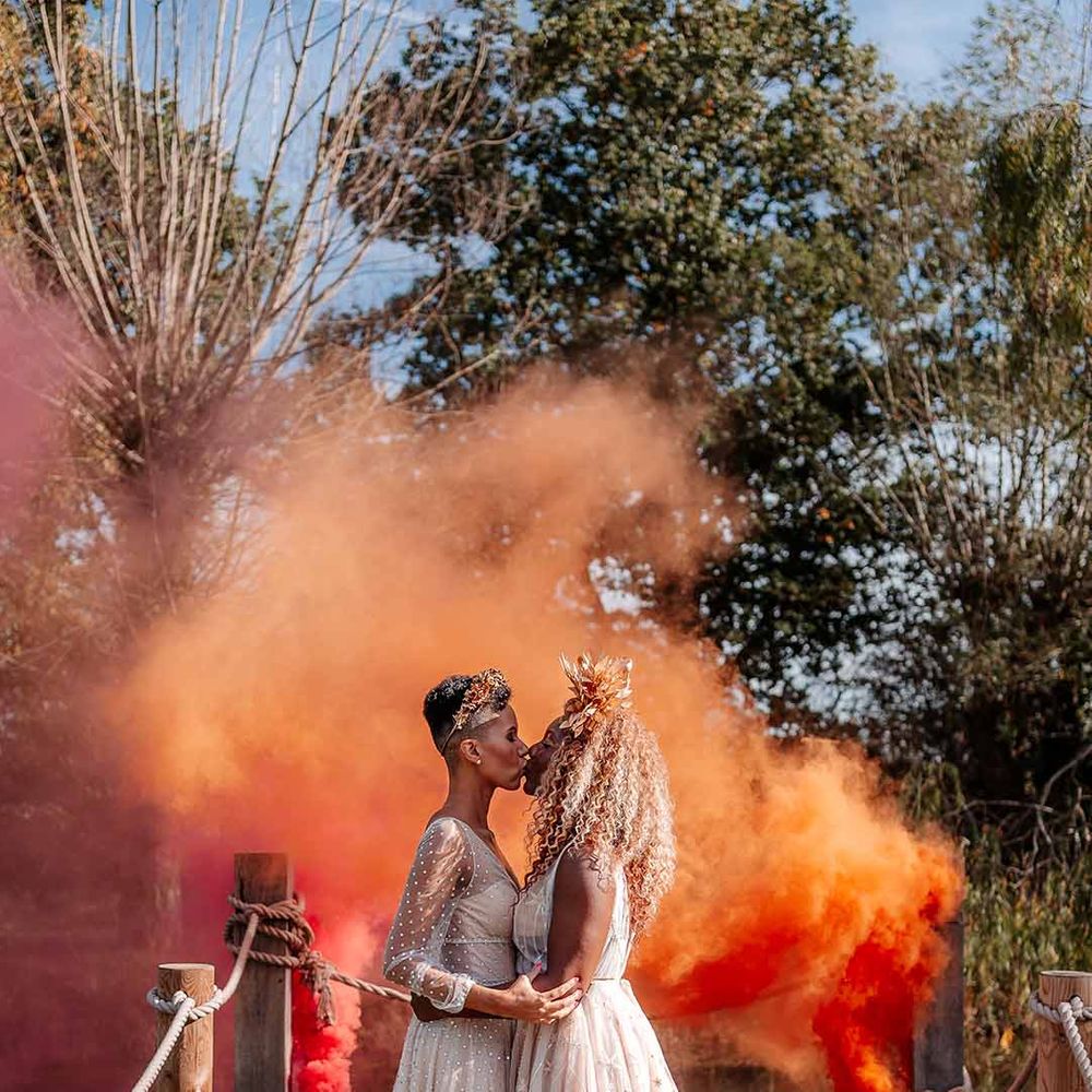 Brides in complimenting boho wedding dresses and golden bridal crowns kissing in front of large orange smoke bomb at Silchester Farm
