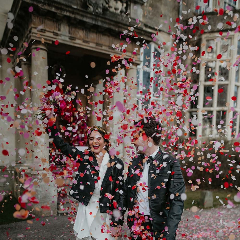Bride and groom in complimenting black personalised leather jackets doing colourful red, magenta and white confetti walk 