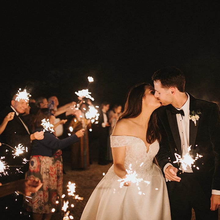 Bride in lace off the shoulder wedding dress kissing the groom as they exit their wedding to sparklers 