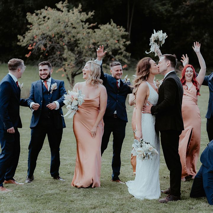 The bride and groom share a kiss as their bridesmaids and groomsmen celebrate and cheer for the kiss 