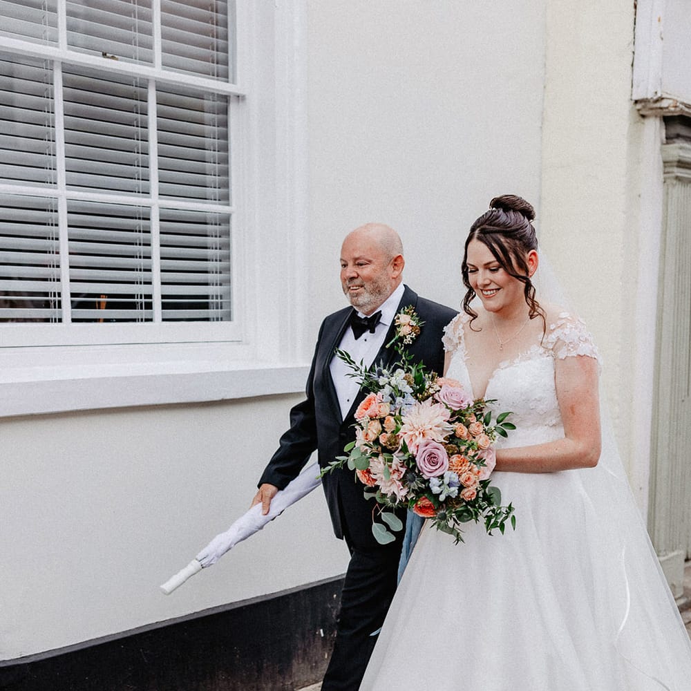 Bride in traditional princess skirt flower Suzanne Neville wedding dress with spring wedding bouquet walking with the father of the bride in black tie 
