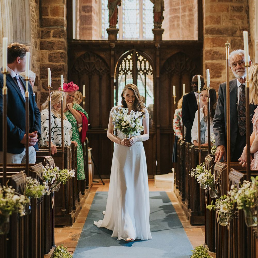 Bridesmaid with short sleeves and bejewelled detail walking down the aisle by herself for the Christian wedding 