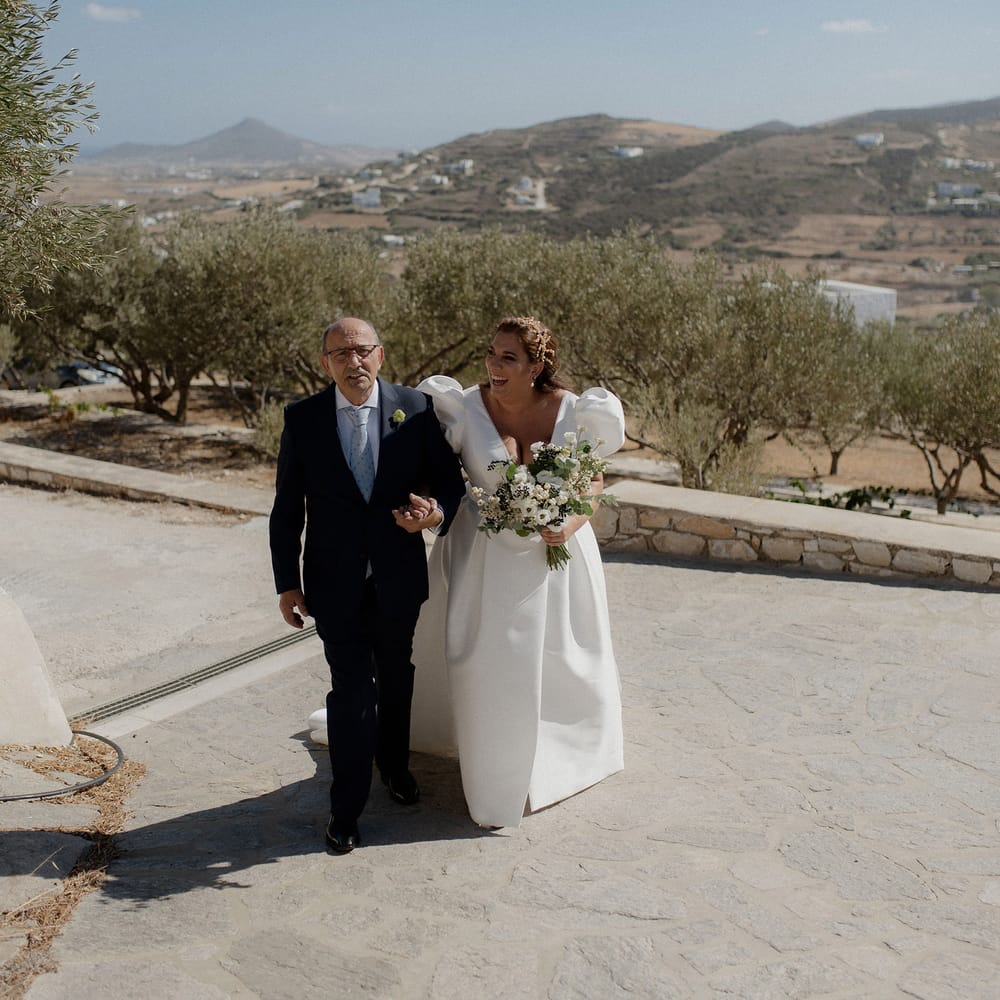 Father of the bride walks bride in satin white dress into wedding ceremony at destination wedding in Paros