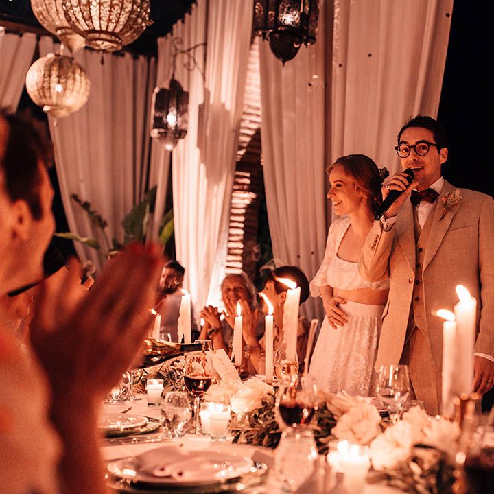 Groom stands up with a microphone to read out his wedding speech with the bride sitting next to him on the top table lit by cosy lanterns 