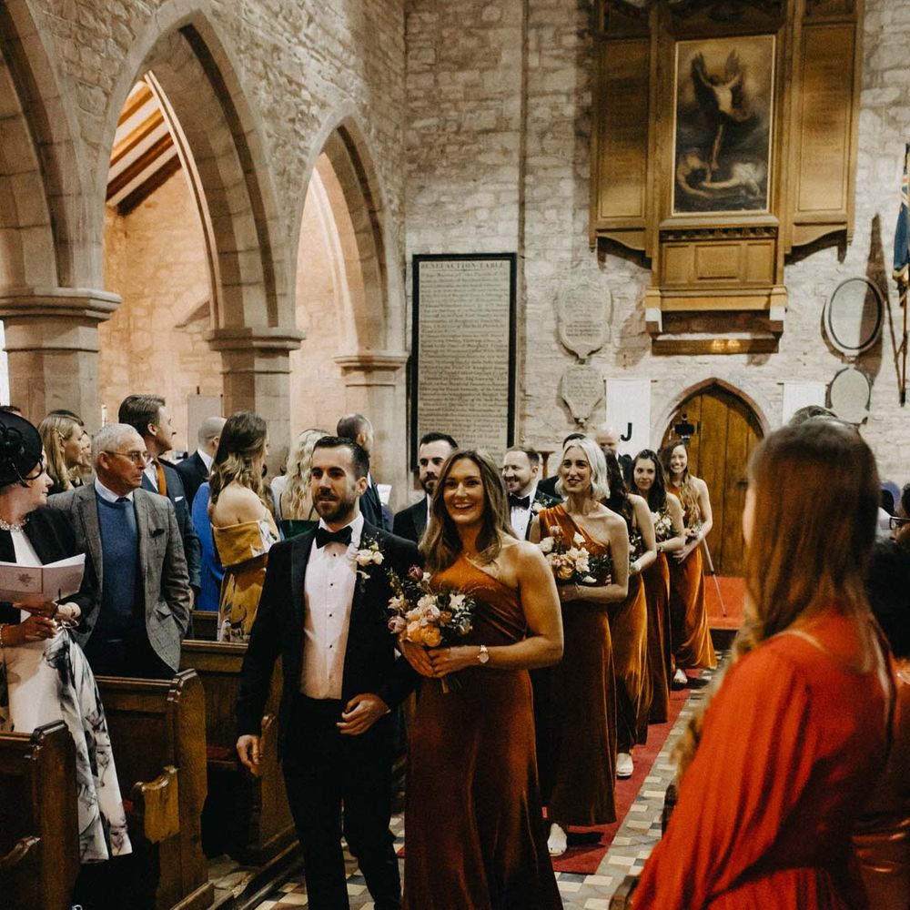 Bridesmaid in one shoulder burnt orange bridesmaid dress walking down the aisle with groom in classic black groomsmen outfit