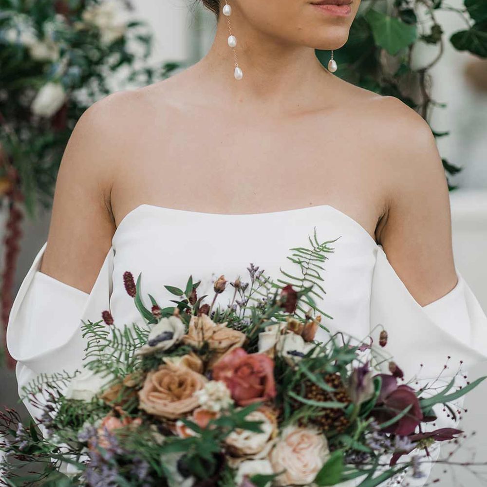 Bride in long sleeve off the shoulder wedding dress with pearl button detailing and a side slit holding dried wildflower bouquet with pearl drop wedding earrings surrounded by suspended wildflower and foliage arrangements in large gold vases at Buxted Park