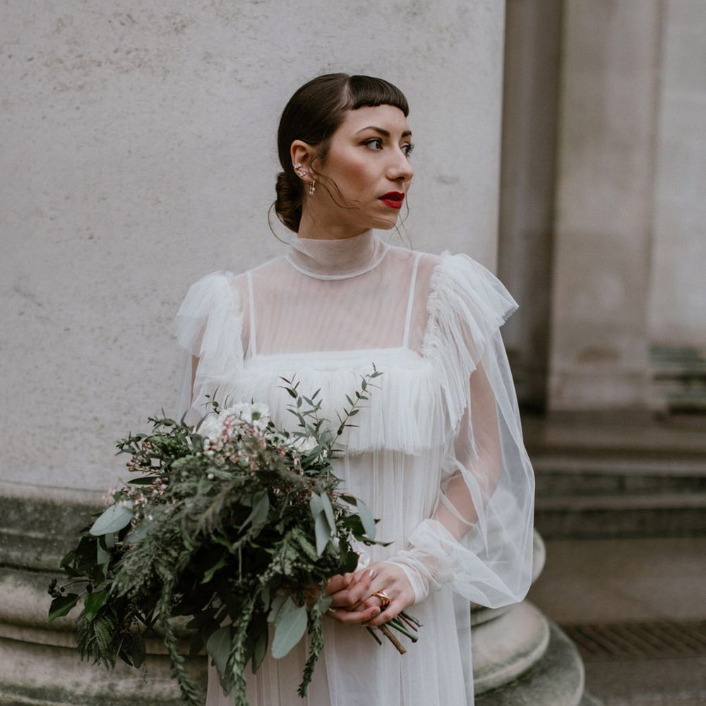 Stylish bride with short fringe and red lipstick wearing a tulle wedding dress with long sleeves and bib detail holding a green and white bouquet 