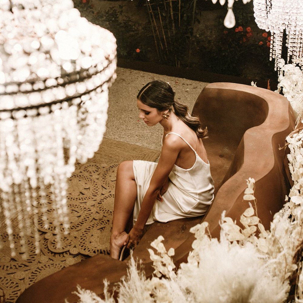 Bride in a fitted satin wedding dress sitting under a canopy of chandeliers and a floor of wicker rugs 