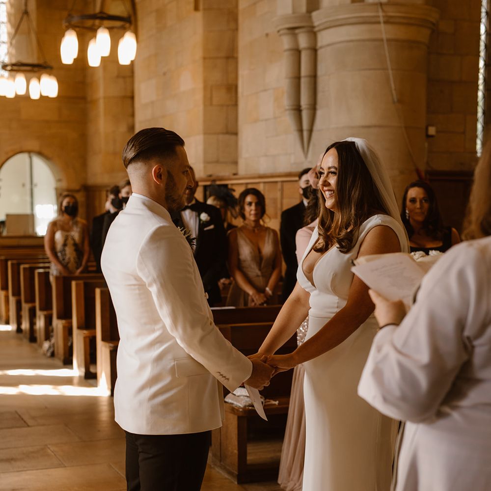Groom in white tuxedo jacket holds hands with bride in white Made With Love wedding dress and white chapel length veil at the church altar