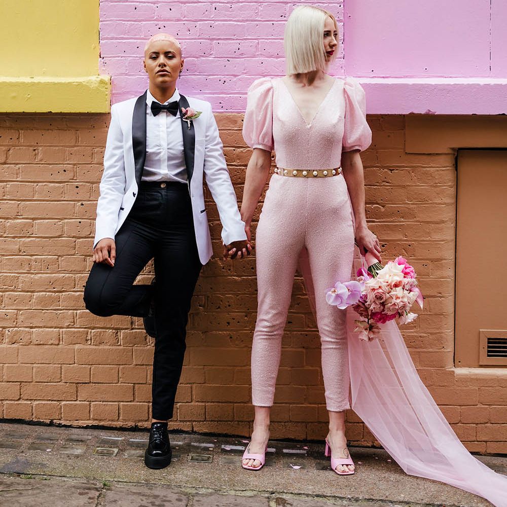 LGBTQ couple in a white tuxedo and pink jumpsuit holding hands in front of a painted wall in Shoreditch 