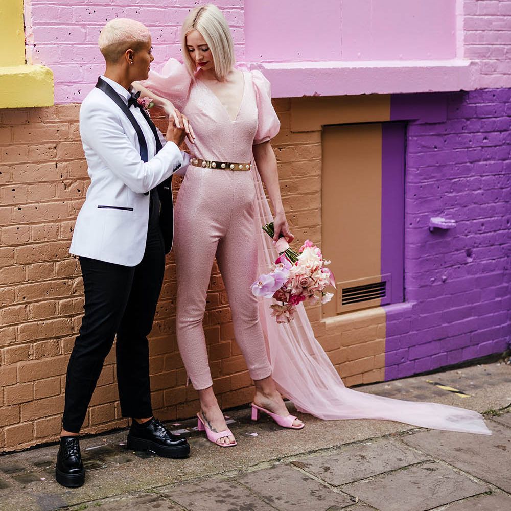Bride in a white tuxedo jacket and platform shoes holding hands with her bride in a pink custom made wedding dress and mules