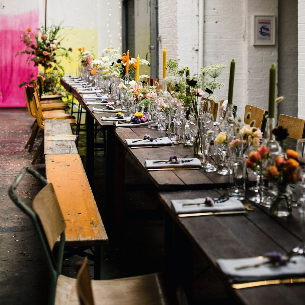 Industrial wedding venue with banquet tables littered with colourful wild flowers in vases and taper candles 