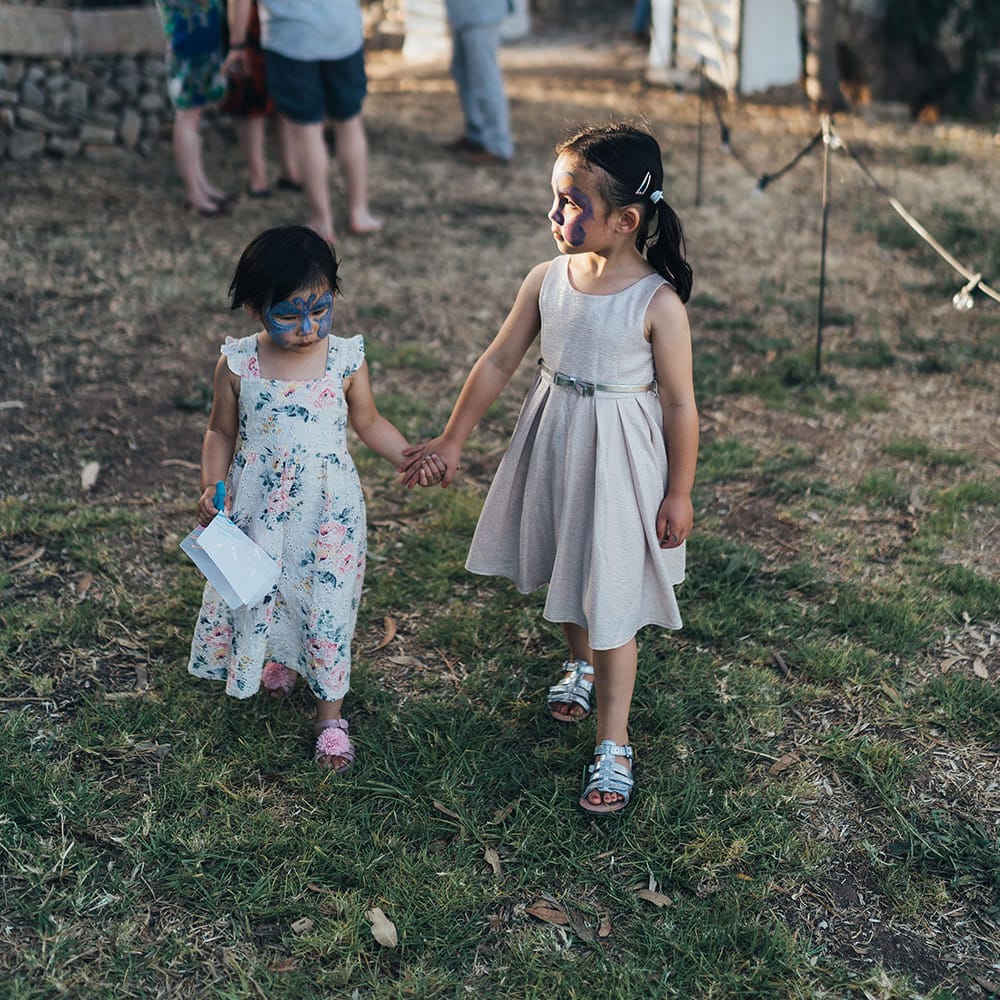 Two little girls walk across grass wearing face paint and dresses whilst holding hands