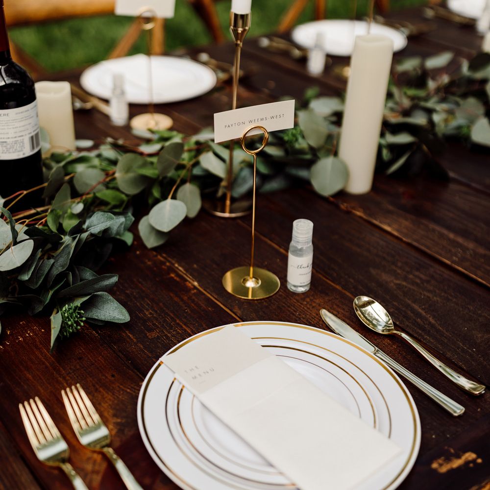White wedding menu resting on white gold rimmed plates all set on wooden wedding table with white candles, and green leaf table garland at backyard wedding