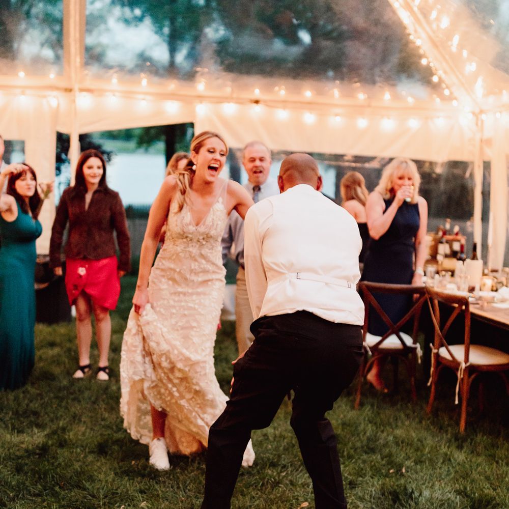 Groom in white waistcoat, black trousers and white Nike Air Force 1 trainers dances in fairy lit marquee as laughing bride in white embroidered Made With Love wedding dress an white Nike Air Force 1 trainers laughs 
