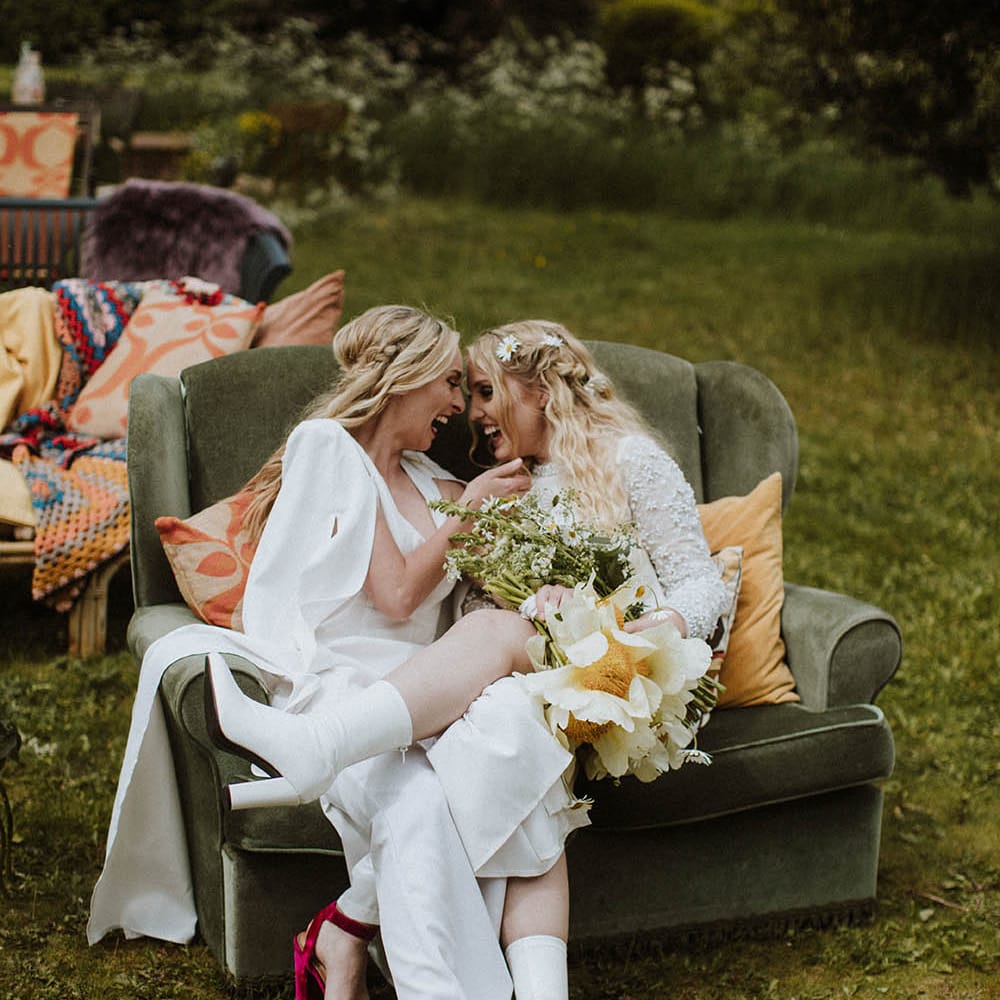 Two brides sitting on a retro sofa at their outdoor sixties wedding 