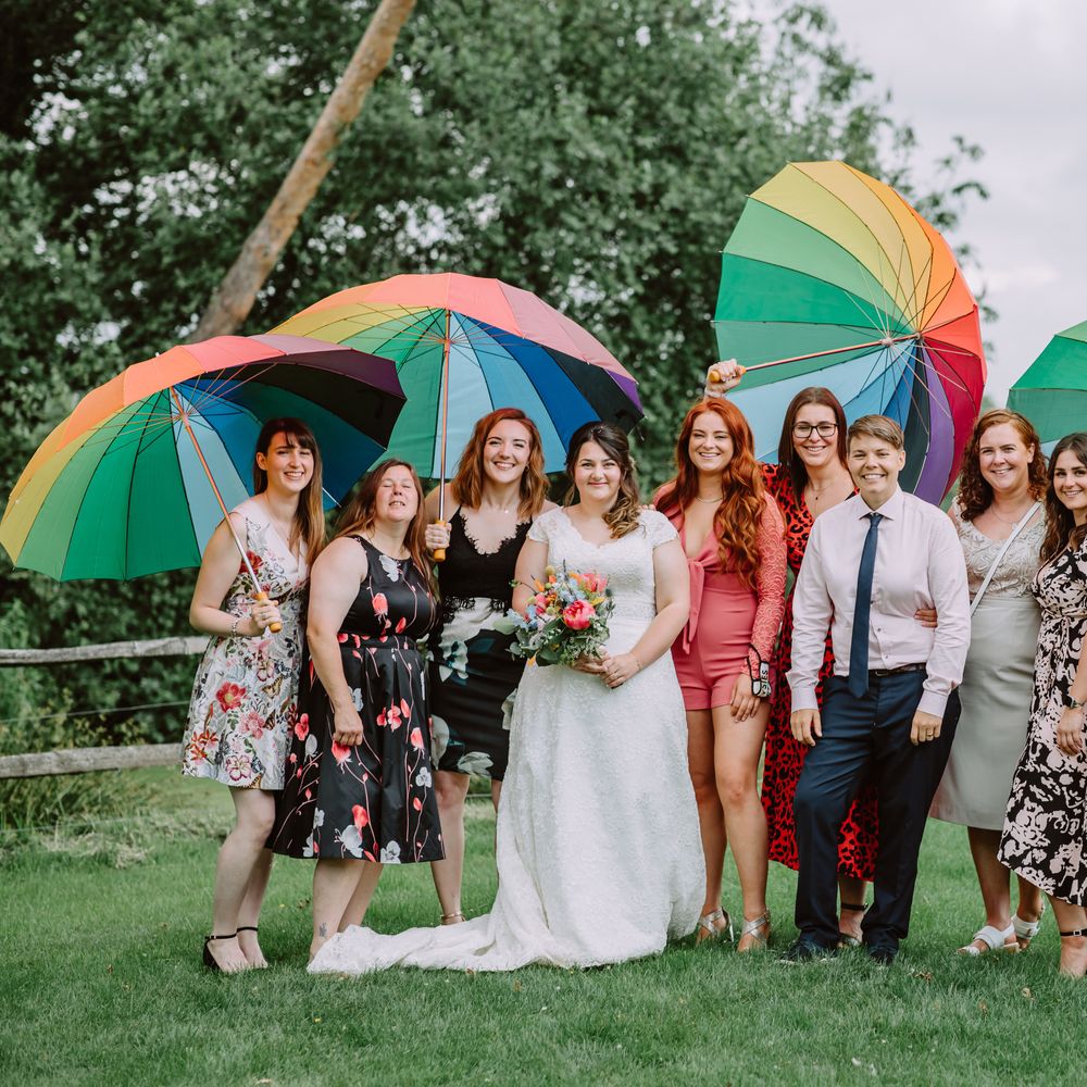 Brides stand with their wedding guests outdoors as they hold Pride umbrellas 