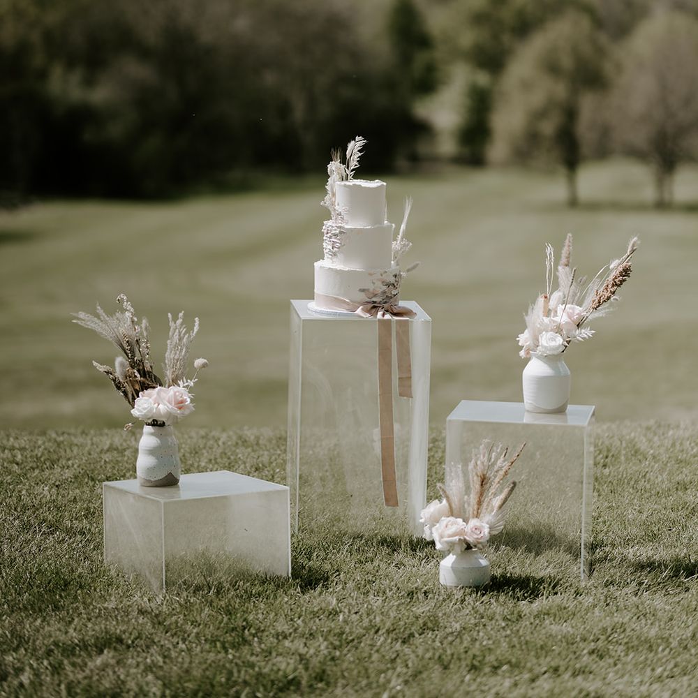 Three tier buttercream wedding cake decorated with dried flowers and grasses resting on an acrylic plinth surrounded by muted flowers in vases 