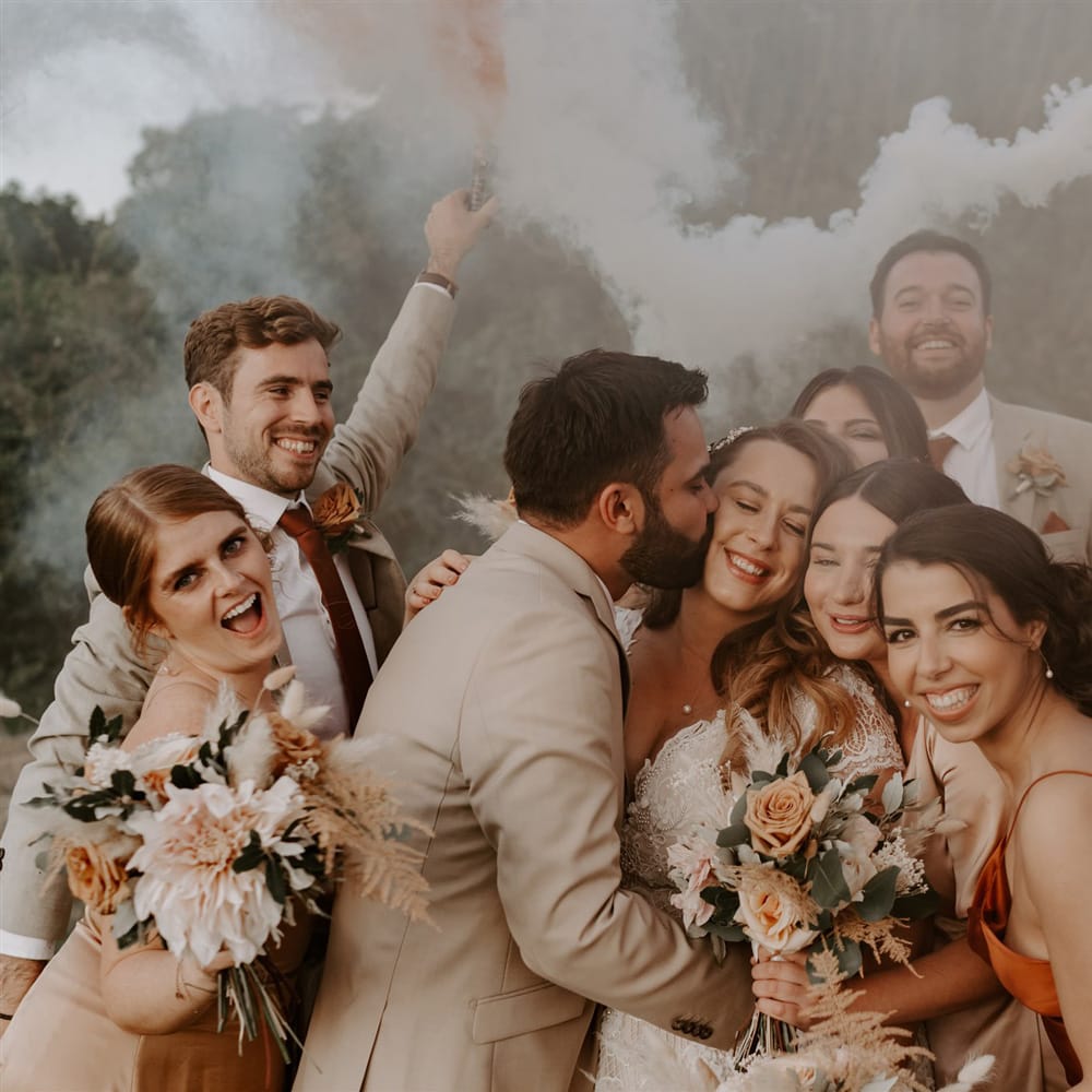 Groom in beige suit kisses cheek of bride in homemade lace wedding dress whilst bridesmaids gather around them and groomsmen smile holding orange and white smoke bombs at outdoor wedding in Bedfordshire