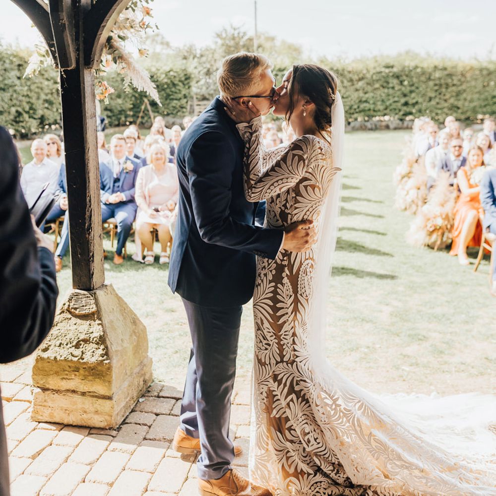 Groom in navy suit and tanned brogues kisses Bride in Rue De Seine (wedding dress) as wedding guests cheer at Barn wedding 