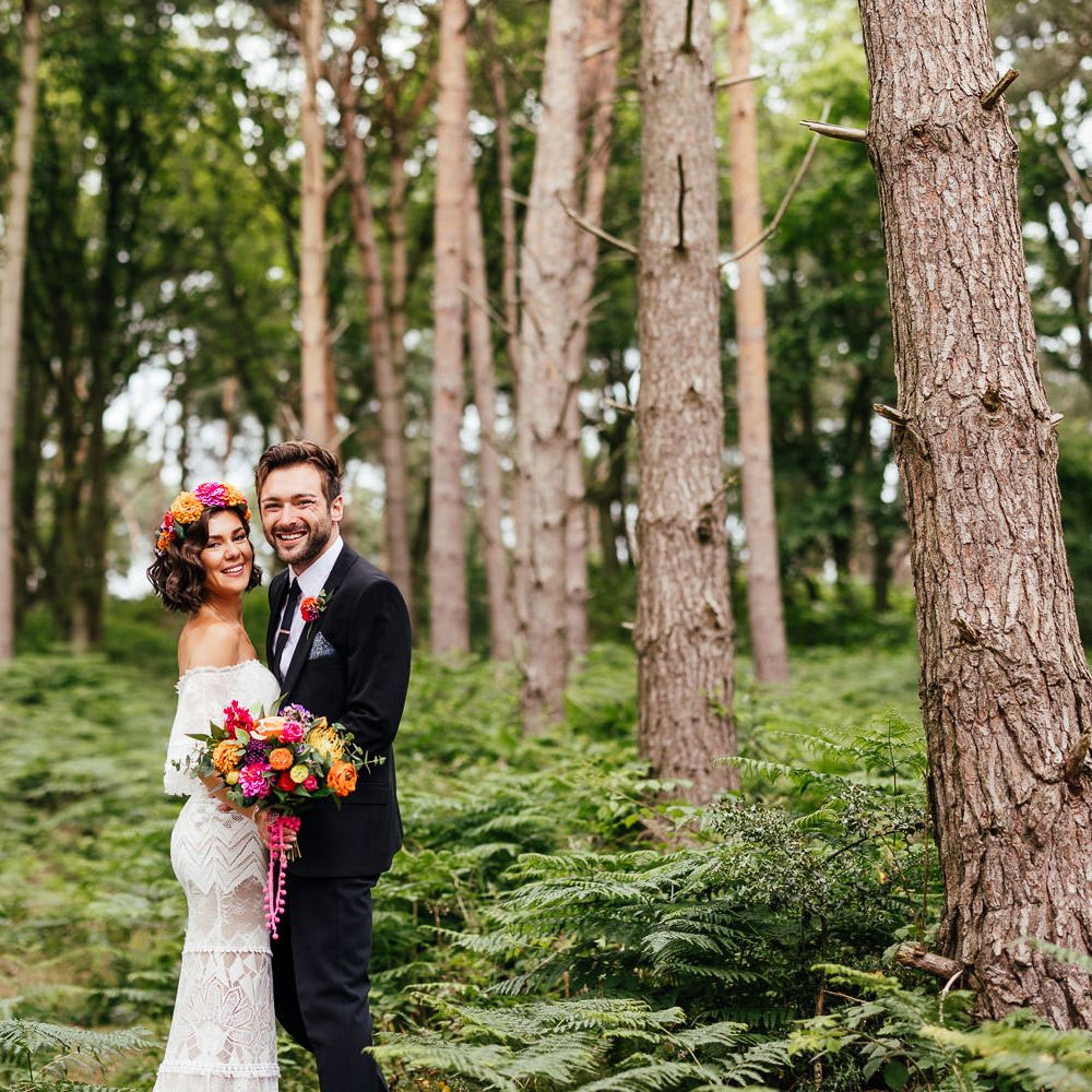 Smiling bride in off the shoulder Grace Loves Lace wedding dress and flower crown holding colourful bridal bouquet stands with smiling groom in black suit in woodland after outdoor wedding ceremony at The Bridal Barn in Claverley