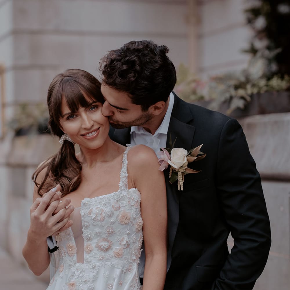 Groom in a black suit with tropical flower buttonhole kissing his brides cheek in an appliqué wedding dress with pockets 