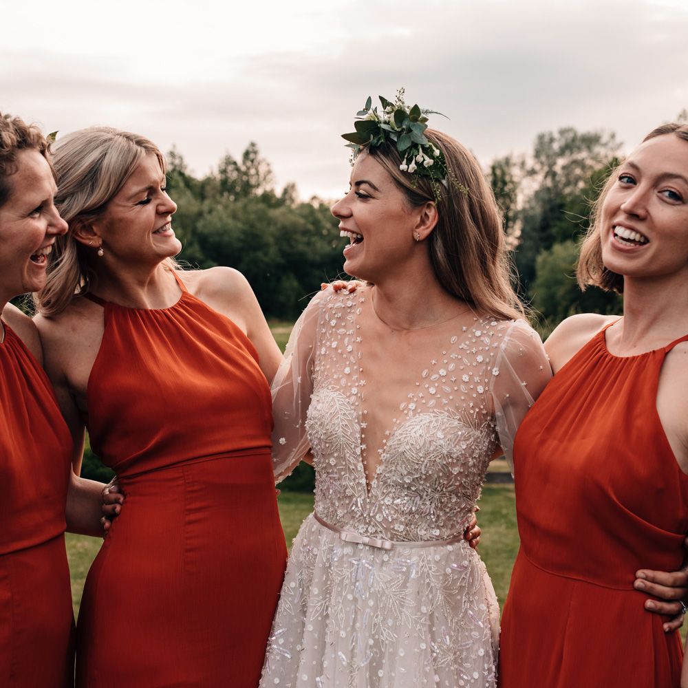 Bride in v neck long sleeve Julie Vino wedding dress and boho flower crown laughs with bridesmaids in red halter neck bridesmaid dresses 