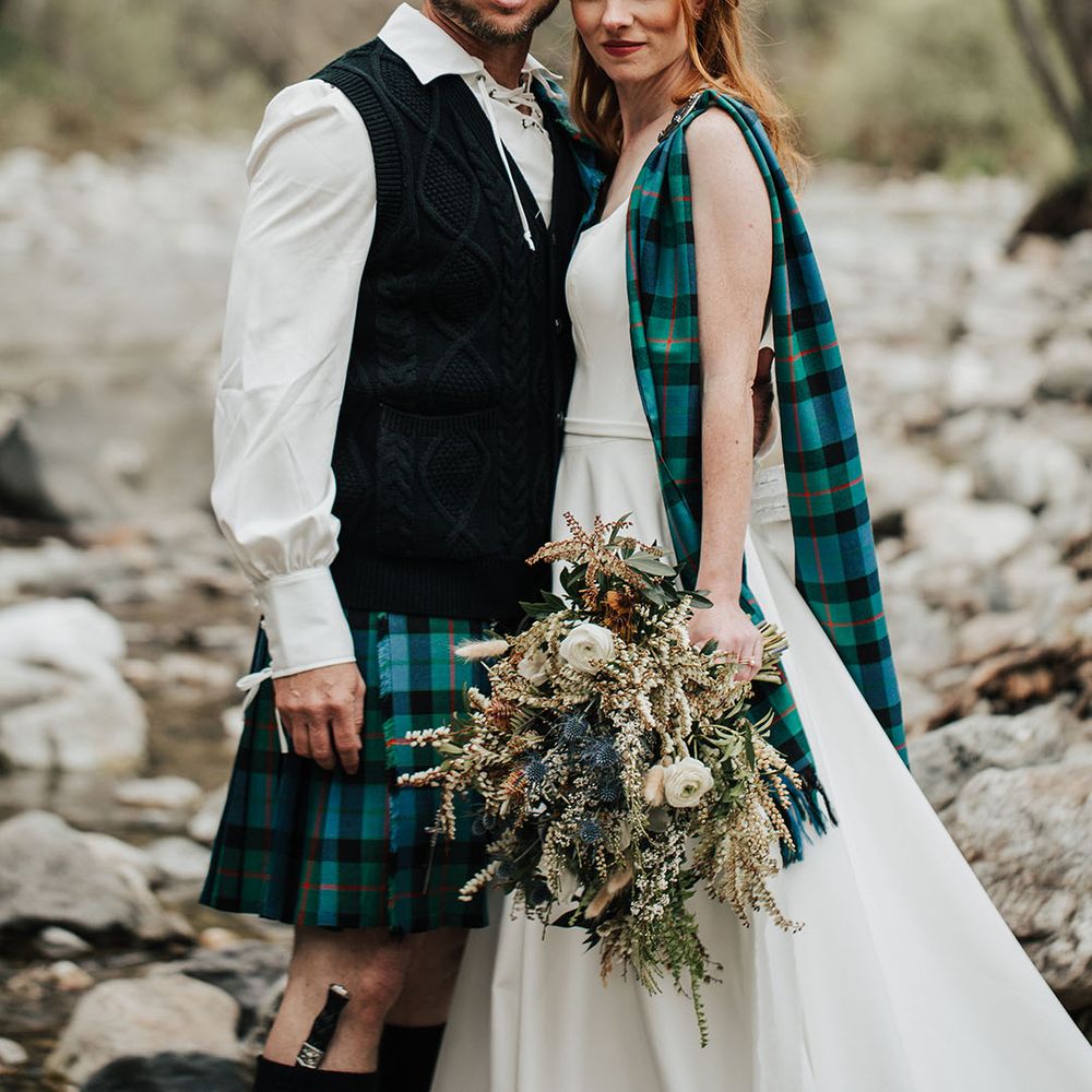 Bride and groom in a tartan kilt and sash standing in a stream holding a boho wedding bouquet with anemones 
