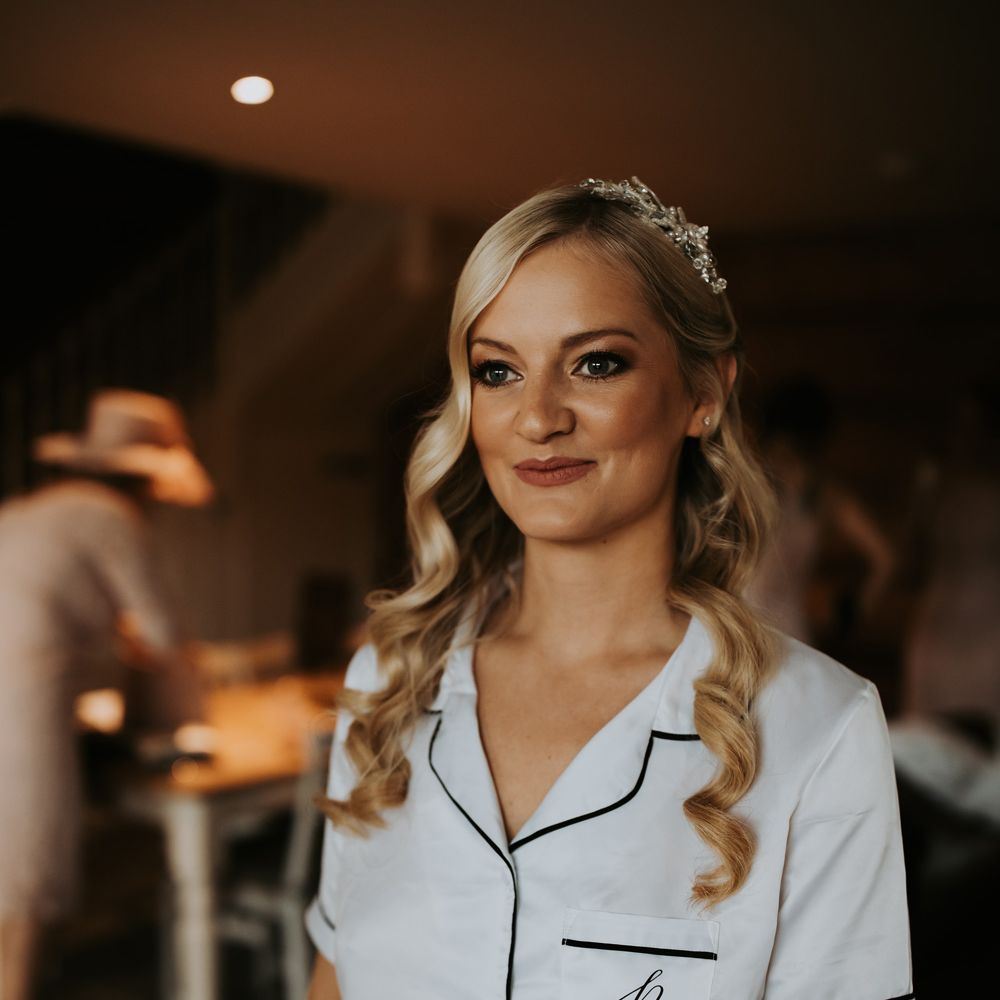 Bride with long curled blonde hair, bridal hair accessory and satin short sleeve bridal pyjamas stands in bedroom before barn wedding