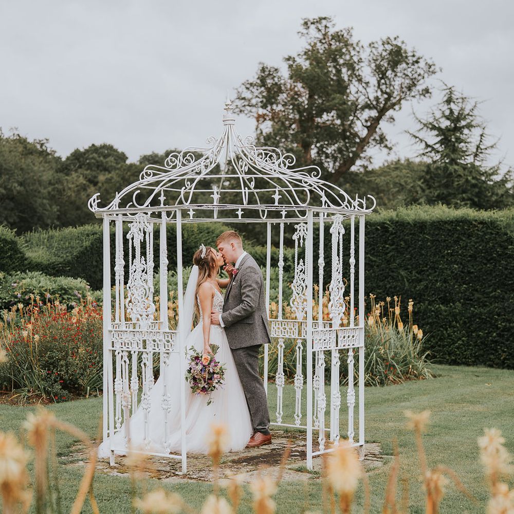 Bride and groom share a kiss in white iron birdcage at rustic country house wedding venue 