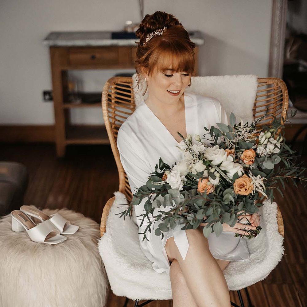 Bride in white satin dressing gown and fluffy socks with satin white mules on fluffy stool with wedding bouquet