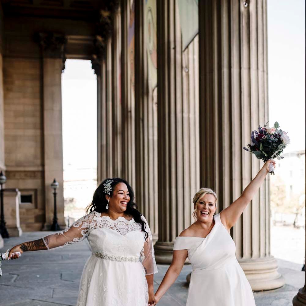 St George's Hall Liverpool wedding with two brides in tea length wedding dresses holding their bouquets in the air 